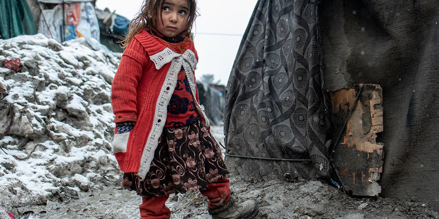 A young Afghan girl is trying her new winter boots outside her tent. She is wearing red trousers, a skirt and a red knit jacket. The ground is wet and snowy.