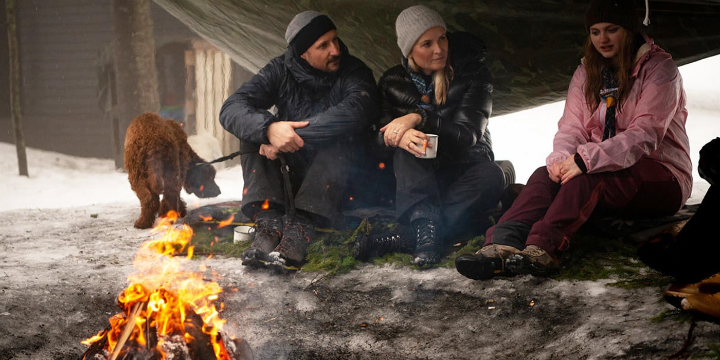 Kronprins Haakon og kronprinsesse Mette-Marit i samtale med en speider. De sitter rundt et bål i en gapahuk.