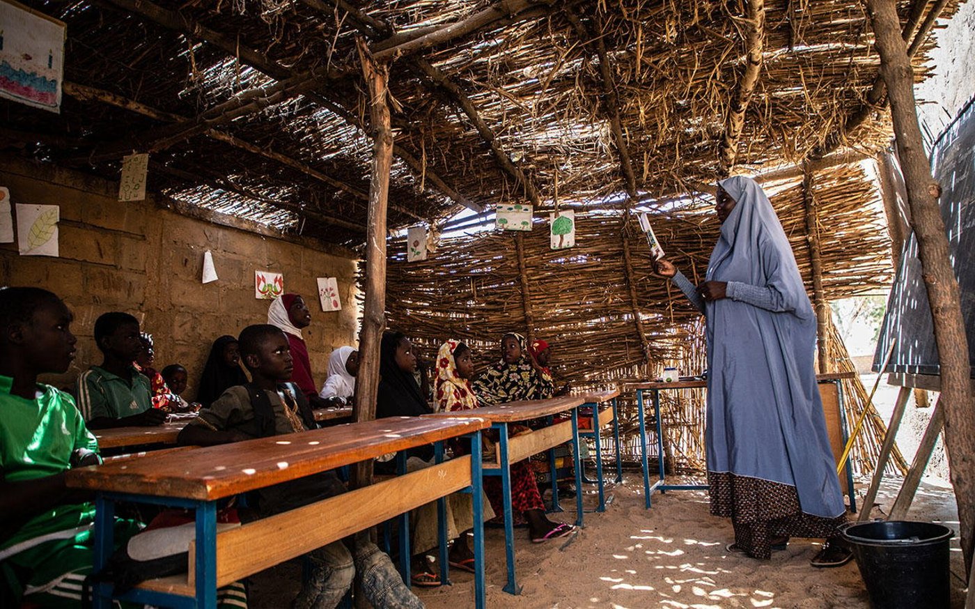 A teacher is standing by a blackboard in front of her class, where children are sitting at their desks.