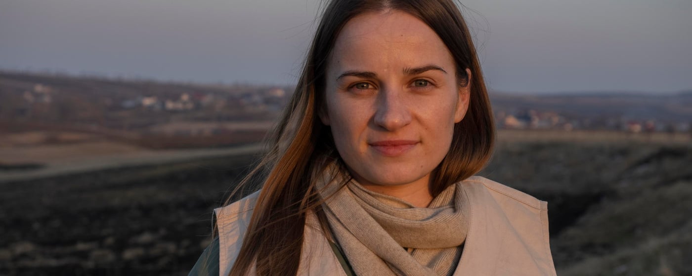 Portrait of Liliia, a woman with long, brown hair. She is wearing a vest with the NRC logo and a grey scarf.