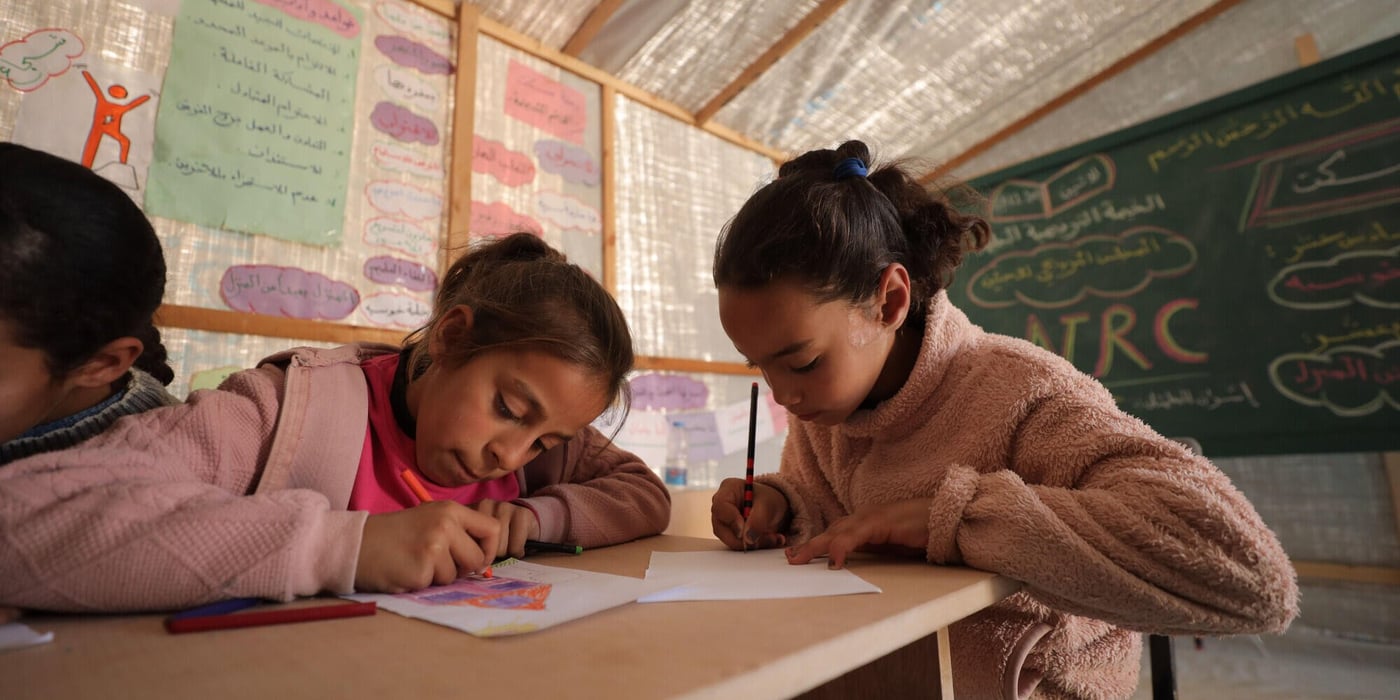 Två flickor sitter vid ett träbord i ett klassrum och ritar eller skriver på papper med färgpennor.