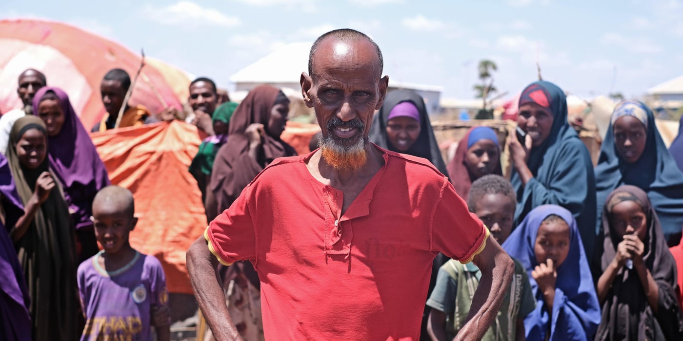 Portrait of a man, looking straight into the camera. He is wearing a red t-shirt, holding his hands on his hips. Behind him is a large group of people.