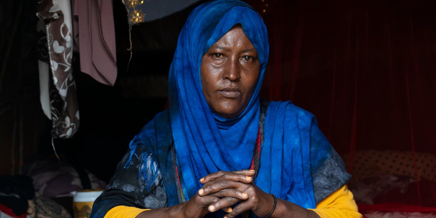 En kvinna sitter inne i ett dunkelt tält och ser rakt in i kameran. Hon bär en klarblå hijab som täcker hår och axlar, och en gul tröja syns under tyget. Händerna är sammanflätade framför henne.