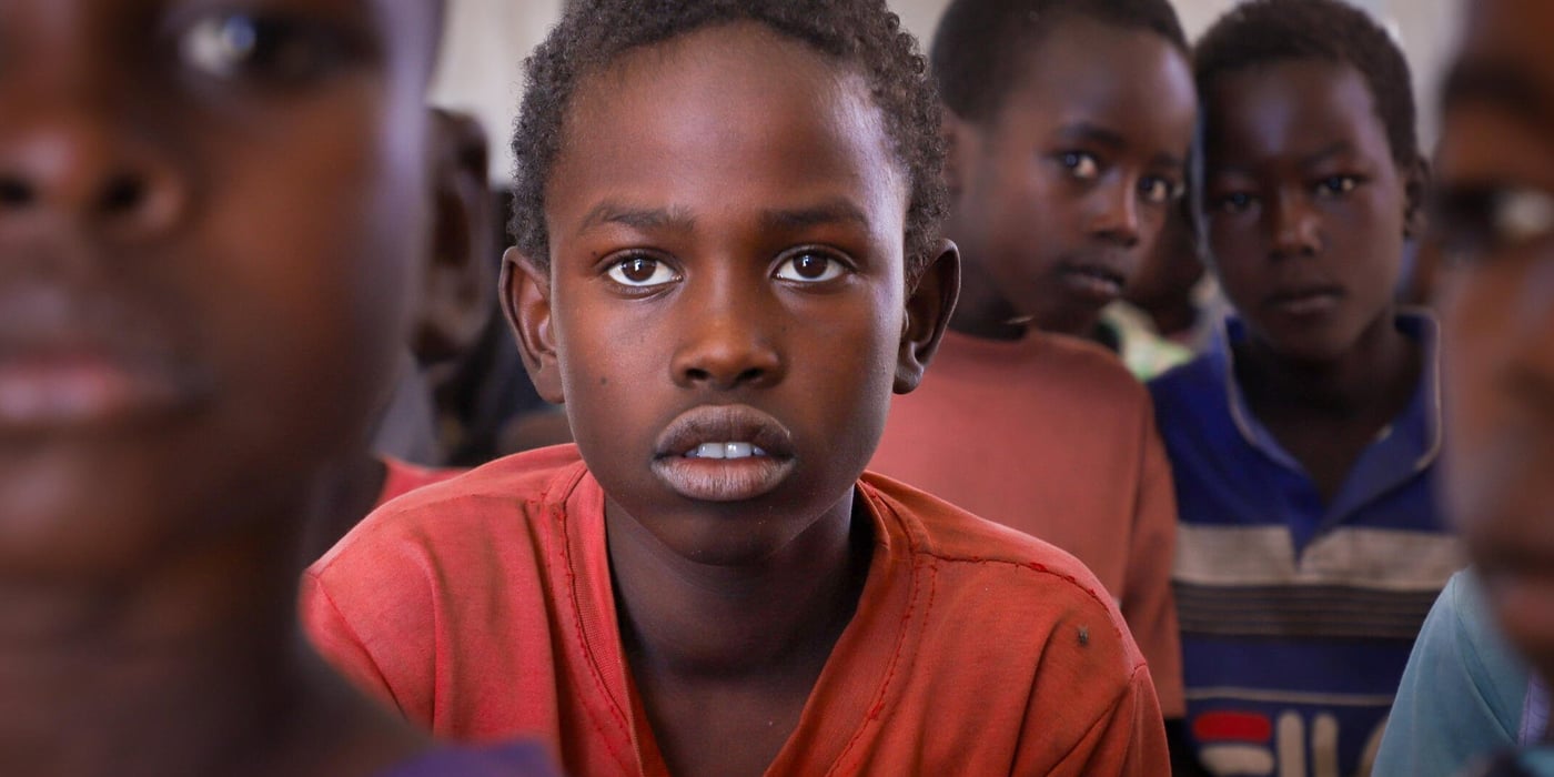 En pojke i röd tröja sitter i ett klassrum med andra barn och tittar in i kameran.