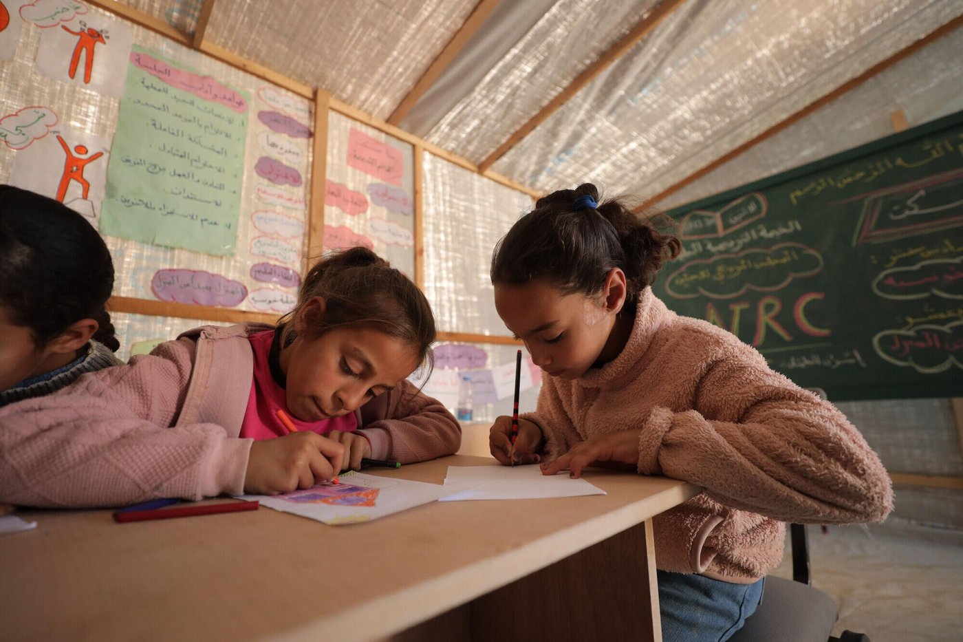 Två flickor sitter vid ett träbord i ett klassrum och ritar eller skriver på papper med färgpennor.