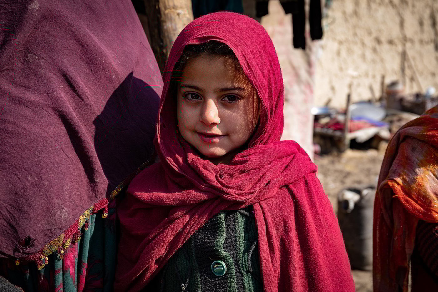 A young Afghan girl wearing a red shawl and a green knit jacket.