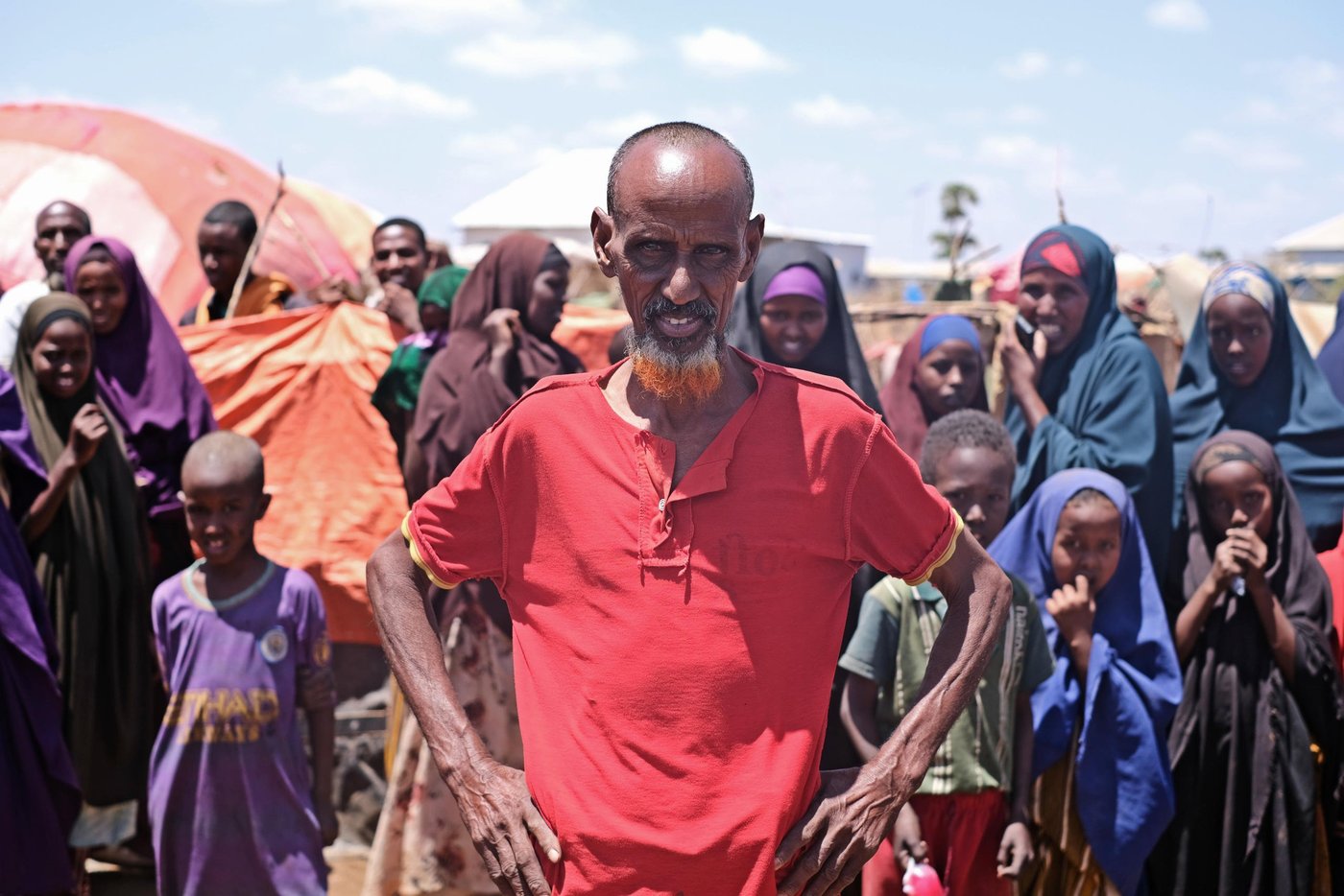 Portrait of a man, looking straight into the camera. He is wearing a red t-shirt, holding his hands on his hips. Behind him is a large group of people.