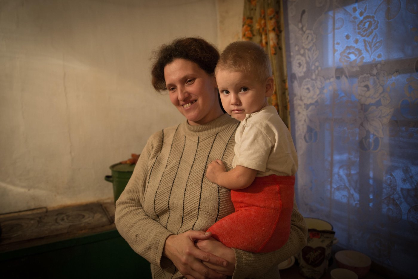 Liliia Poturoieva is 39. She has experienced terrible events that completely changed the life of herself and her large family and left an indelible mark on everyone's soul. She lives in the frontline village of Verkhnia Vilkhova in Stanytsia Luhanska district, Luhansk region.
Liliia has six children. The oldest is Oleksandr. He is 20. He lives separately from his parents and earns his own living. Ihor is 12. He is a shy and calm schoolboy, always ready to help parents with the housework. Her eldest daughter Iryna is 10. She is a schoolgirl and mother's main housekeeper. She cooks and looks after younger children. Yana is 7. She is a first-grade pupil. She is a cheerful, active and very energetic girl. The children are forced to go to school in a neighboring village 5 kilometers away, since there is simply no school in their village.
Her youngest son Illia,5, and daughter Aryna, 2, are always staying at home with their mother. The family cannot afford a kindergarten for children. Now Liliia is pregnant again. This will be the seventh child in the family. Her husband Viktor helps her to cope with all of them.
Before the conflict, the whole big family lived in a small house with two rooms. There was no work. The main income was child allowances and random earnings from the cows, goats, rabbits and poultry.
“There was no work in the village. There was no opportunity to travel to other settlements to work. We tried to make money by selling milk”.
In February 2015, at midnight, heavy shelling began. Viktor, Liliia's husband, went out into the yard to see the direction of the shots. Suddenly a shell fell near their yard, and the second shell landed 7 meters away from Viktor, who was standing on the house porch. Viktor got a serious shell shock and lost his hearing for a while.
The blast wave knocked out all the windows in the house. Under one of the windows there was a child's bed, where Liliia’s youngest son slept. That night Liliia saved the life of her son with her son.
“When the blast hit, I covered my baby with my own body, and the glass from the broken window hit me in the back”.
The consequences of that shelling are still noticeable. Liliia has problems with her speech. She is stuttering. She is not the only one.
“The children experience anxiety, concentration problems, fears and, as a result, urinary incontinence”.
The house was seriously damaged in the blast. All the windows were broken, the front walls were destroyed and the roof and the foundation were seriously damaged. However, Liliia and her family did not dare to leave because it was too risky to leave the house unattended. They tried to repair the house on their own. They covered the broken windows with plastic wrap. They even had to spend their nights in the basement because it was the warmest and the safest place in their household.There was no help from the local authorities.
“At first I didn’t want to ask for help. I did not believe that anyone would really help us. My husband advised me to go to the NRC office and find out about the Heavy repair project, because he heard that NRC had already helped someone in the village”.
Now the house is in the process of restoration. There is a new slate on the roof. Renovation works on the walls have been completed. Three small rooms for the children have been completed. New windows have been installed and internal works are being carried out now.
“The main thing for me now is that the active phase of the war does not break out again. I am afraid that my house will be destroyed again. After all, we hear the sounds of shots every night”, Liliia says.
Short version: Liliia Poturoieva, 39, mother of six, soon seven children lives close to the contact line in eastern Ukraine. At midnight, February 2015, another round of heavy shelling began and hit Liliia’s house. That night she saved the life of her youngest son. “The blast wave knocked out all the windows in the house, but I covered my baby who slept in his bed right under the window with my own body and the glass pieces hit me in the back”. After that night Liliia began having problems with her speech. The stuttering is a daily reminder of the war.
Photo: Ingebjørg Kårstad/Norwegian Refugee Council