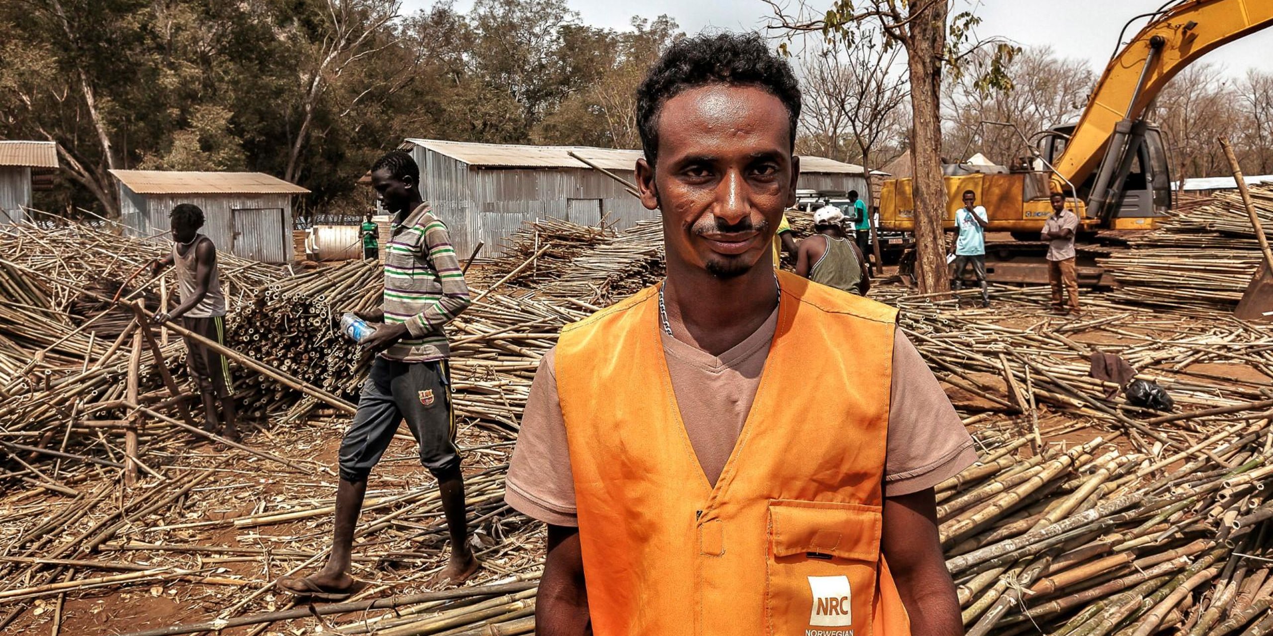 Hintsa Gebreziabher in an orange vest with the NRC logo on a pocket. Behind him are lots of people, building materials and a large excavator.