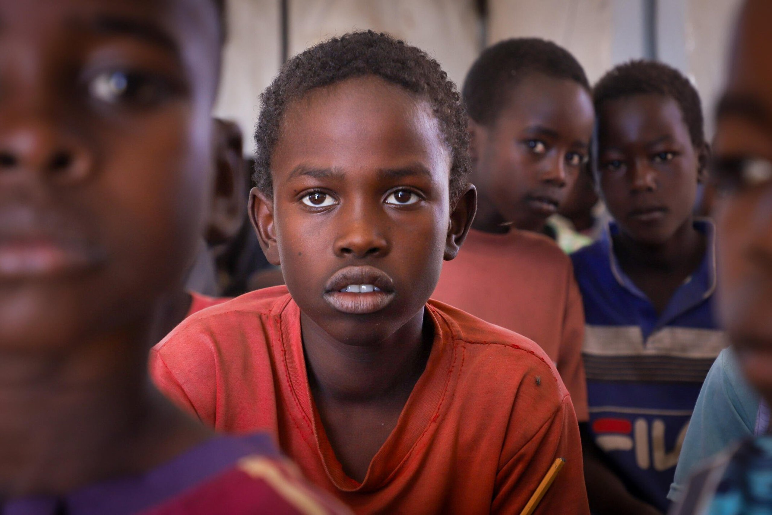 En pojke i röd tröja sitter i ett klassrum med andra barn och tittar in i kameran.