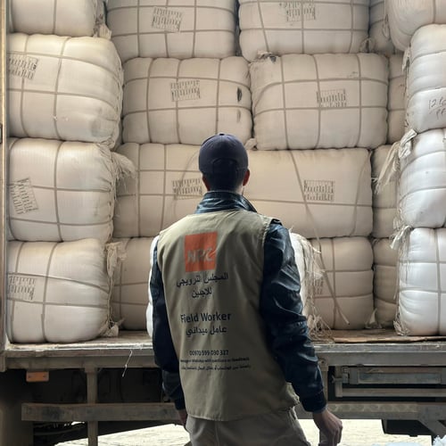 En man står bakom en lastbil lastad med stora vita säckar, staplade tätt intill varandra. Han bär en väst märkt med Norwegian Refugee Council och texten – Field Worker – och ser ut att inspektera eller förbereda lossning av humanitärt bistånd.