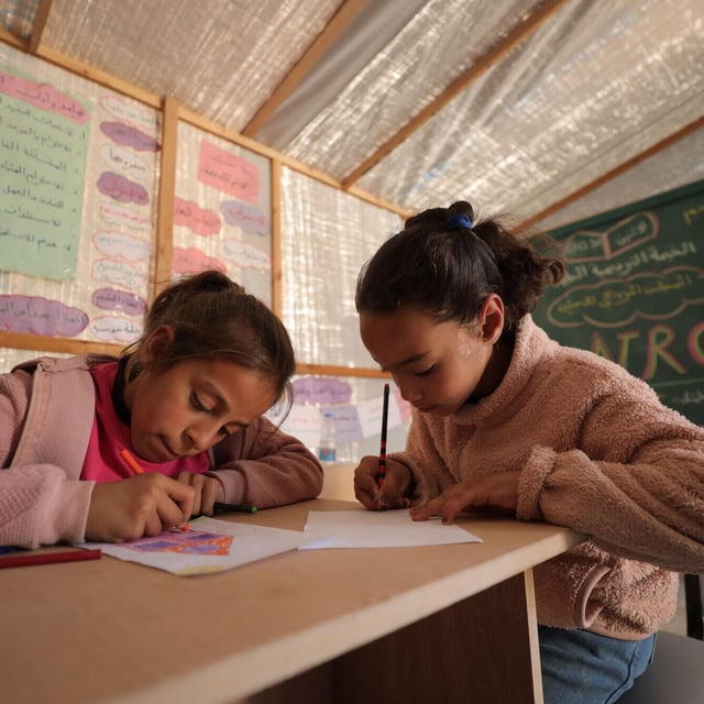 Två flickor sitter vid ett träbord i ett klassrum och ritar eller skriver på papper med färgpennor.