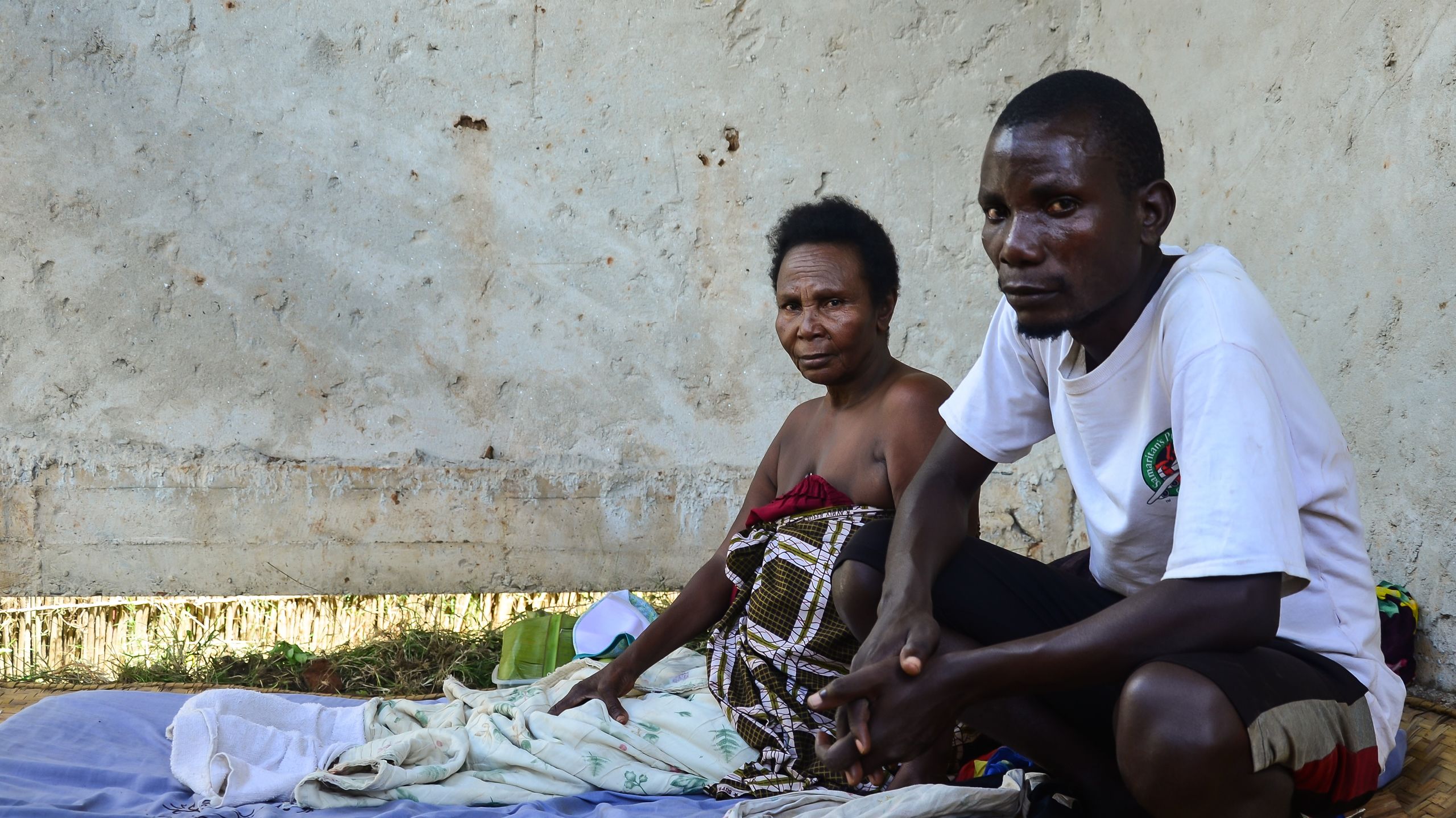 En mann og en kvine sitter på bakken foran en gråhvit murvegg.