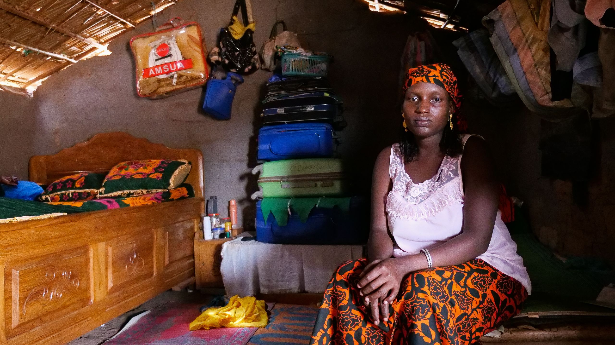 En kvinne sitter inne i huslyet sitt. Hun har på seg en hvit overdel, et oransje sjal på hodet, og store øredobber.