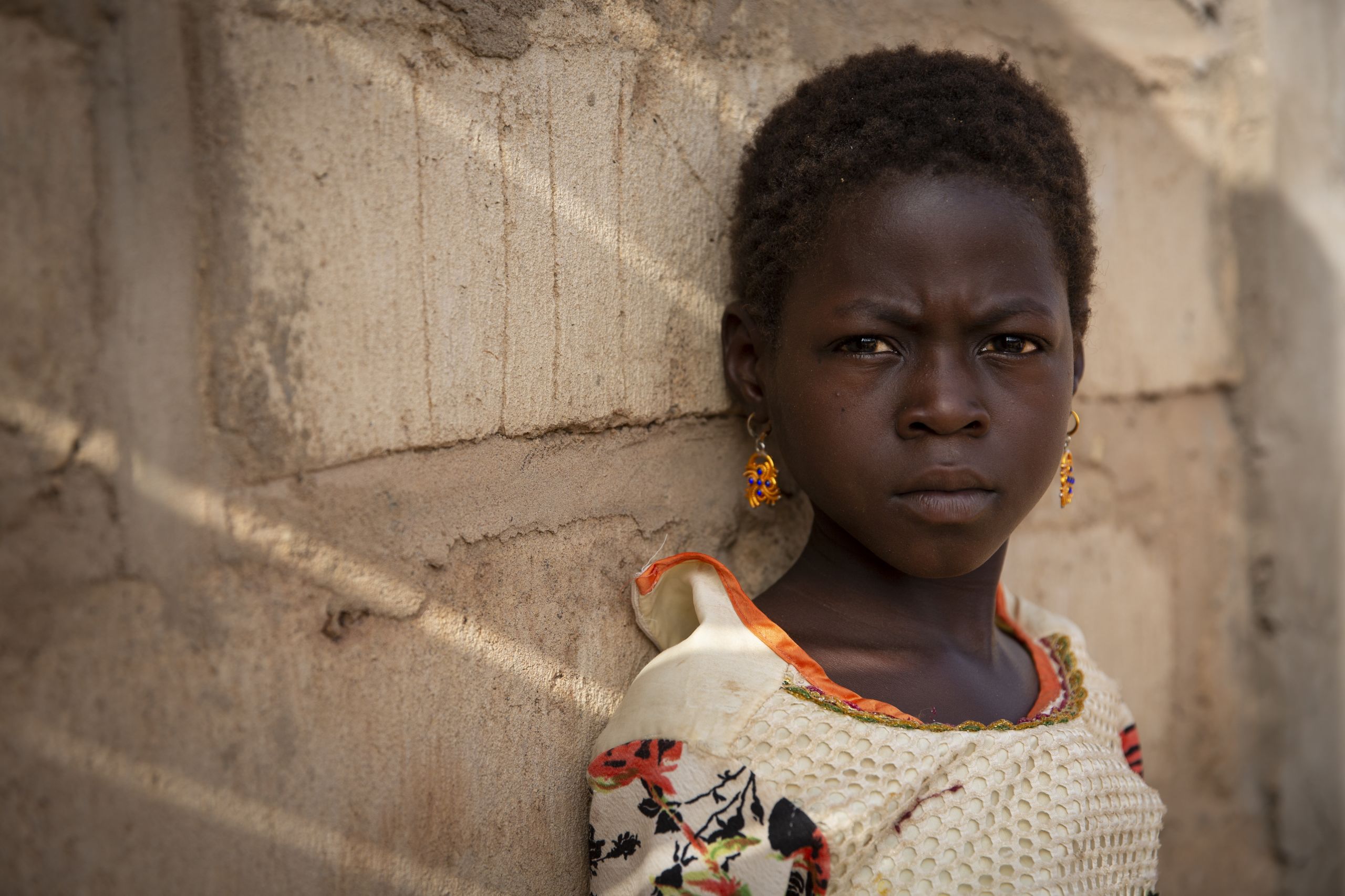 Portrett av Nafi. Hun har et alvorlig blikk og ser rett i kameraet, mens hun lener seg inntil en murvegg. Hun har store, oransje øredobber og en hvit overdel med oransje detaljer.