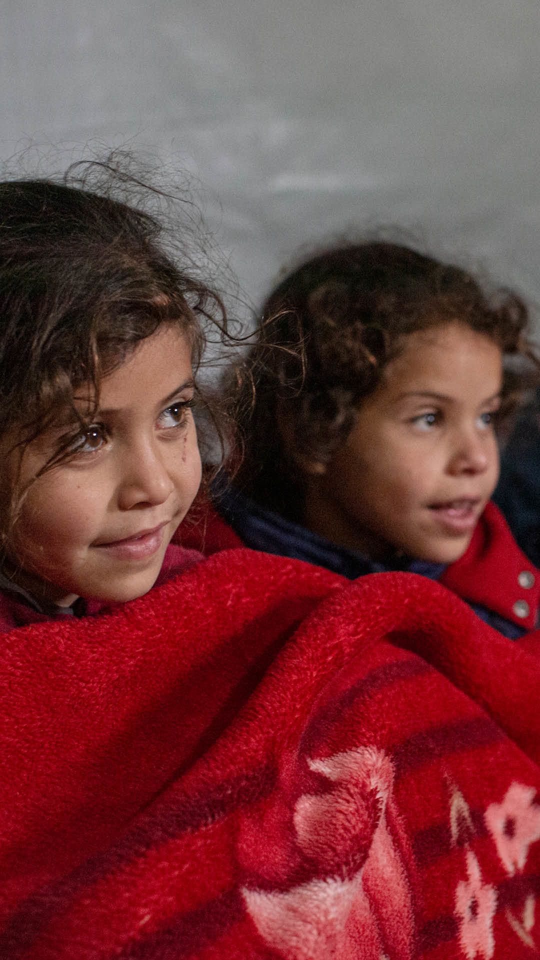 Amira (8) og Aya (6) sitter under et rødt teppe i teltet de bor i.