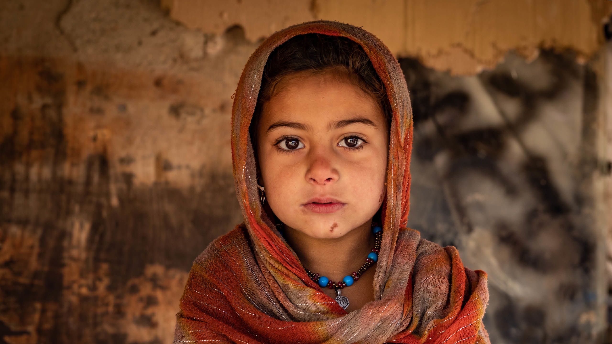 Amina ser rett i kameraet. Hun har et oransje sjal over hodet og et blått smykke rundt halsen.