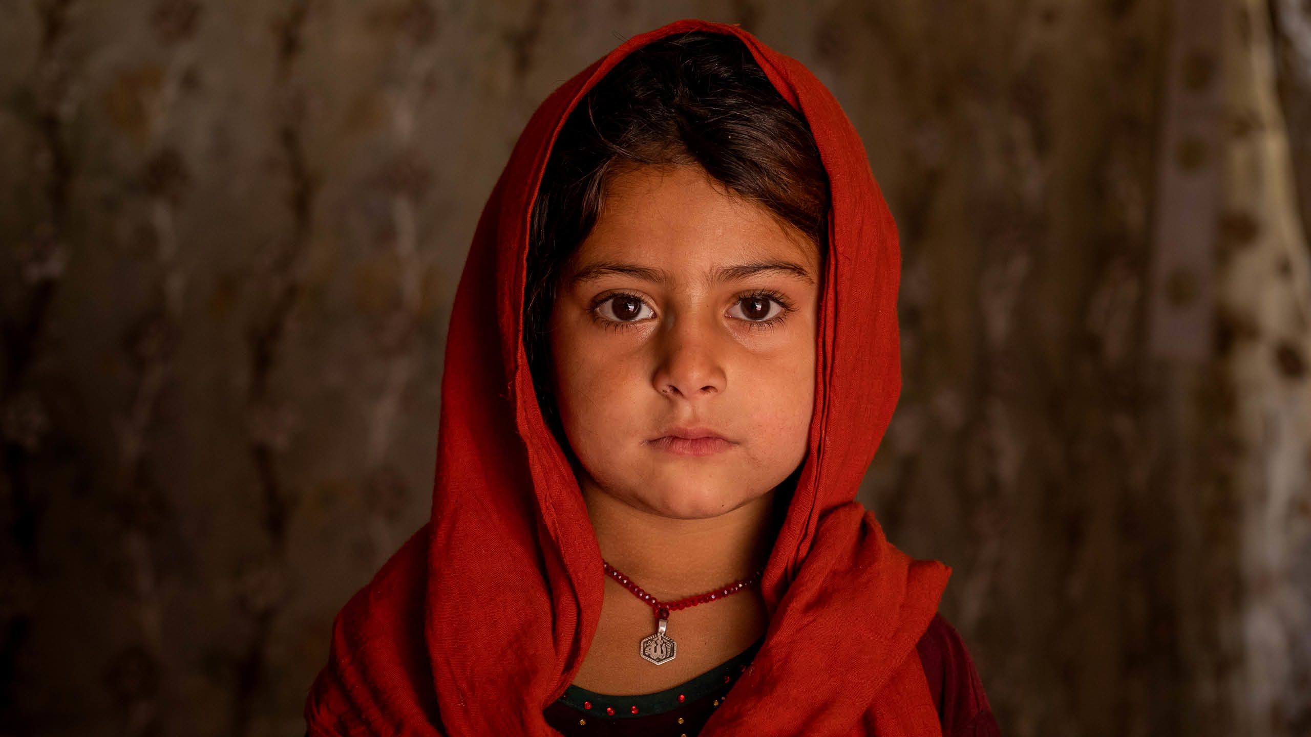Amina (7) ser rett i kameraet. Hun har et rødt sjal over hodet og et rødt smykke rundt halsen.
