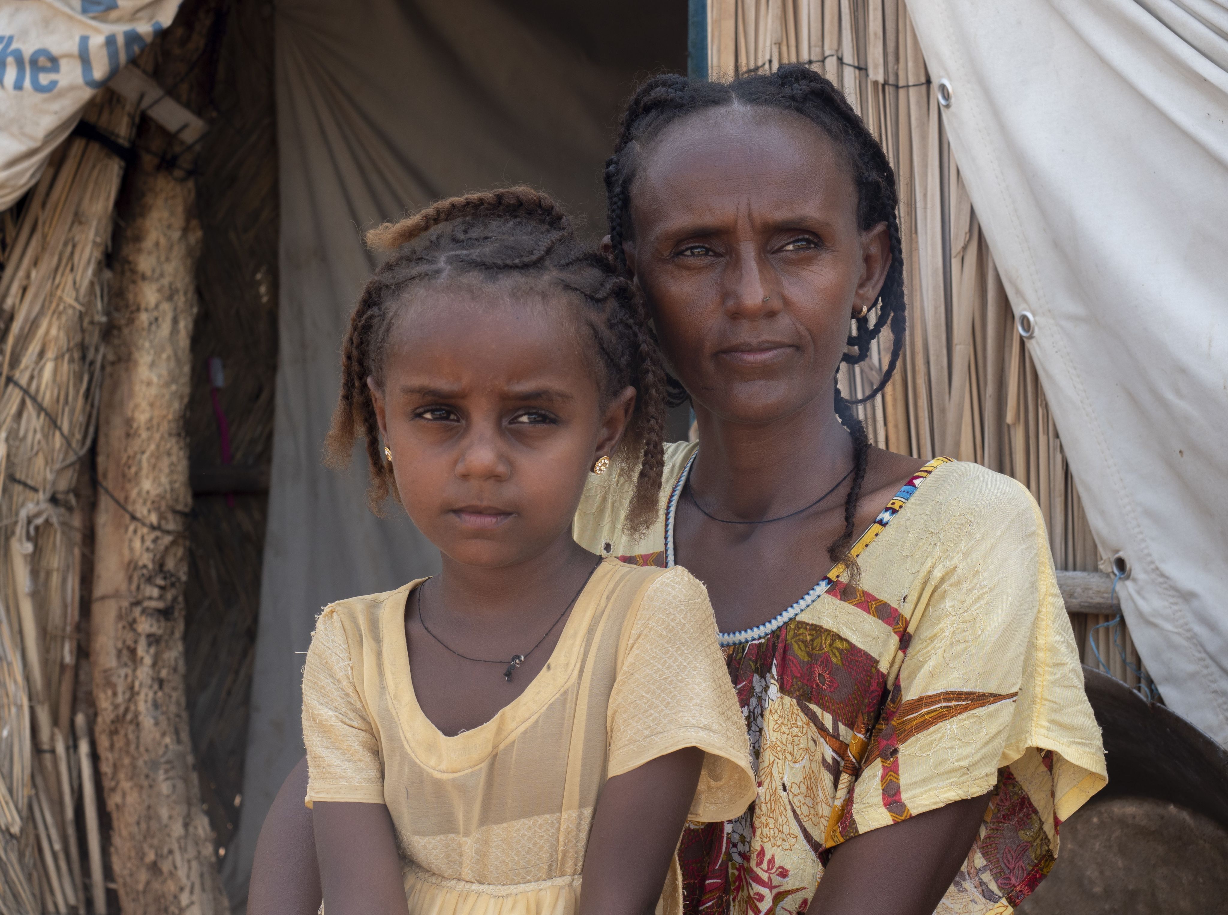 A mother and her daughter, both dressed in yellow, are sitting together in front of their home.