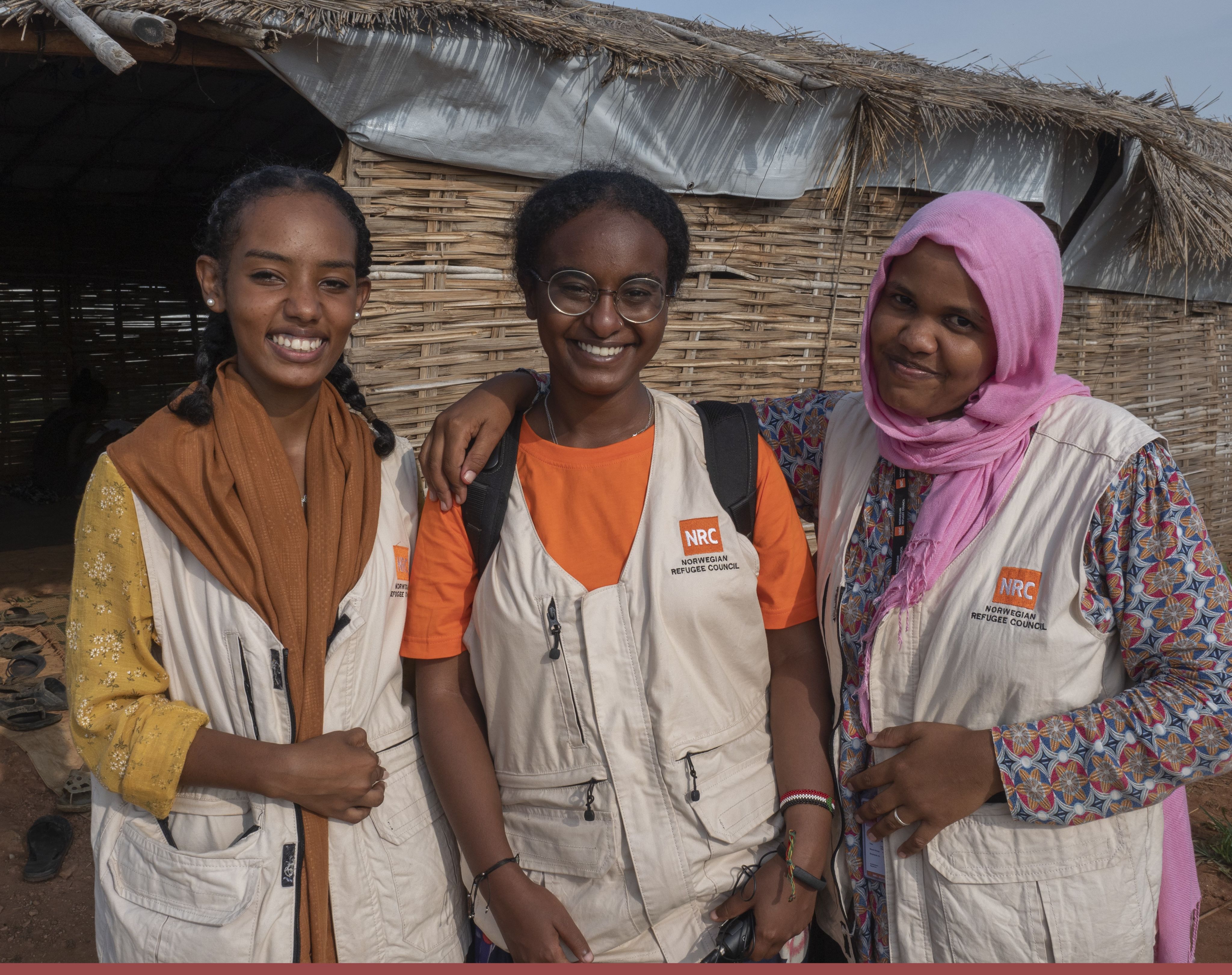 Three NRC aid workers smile for the camera. They are all wearing cream-coloured vests bearing the Norwegian Refugee Council's logo.