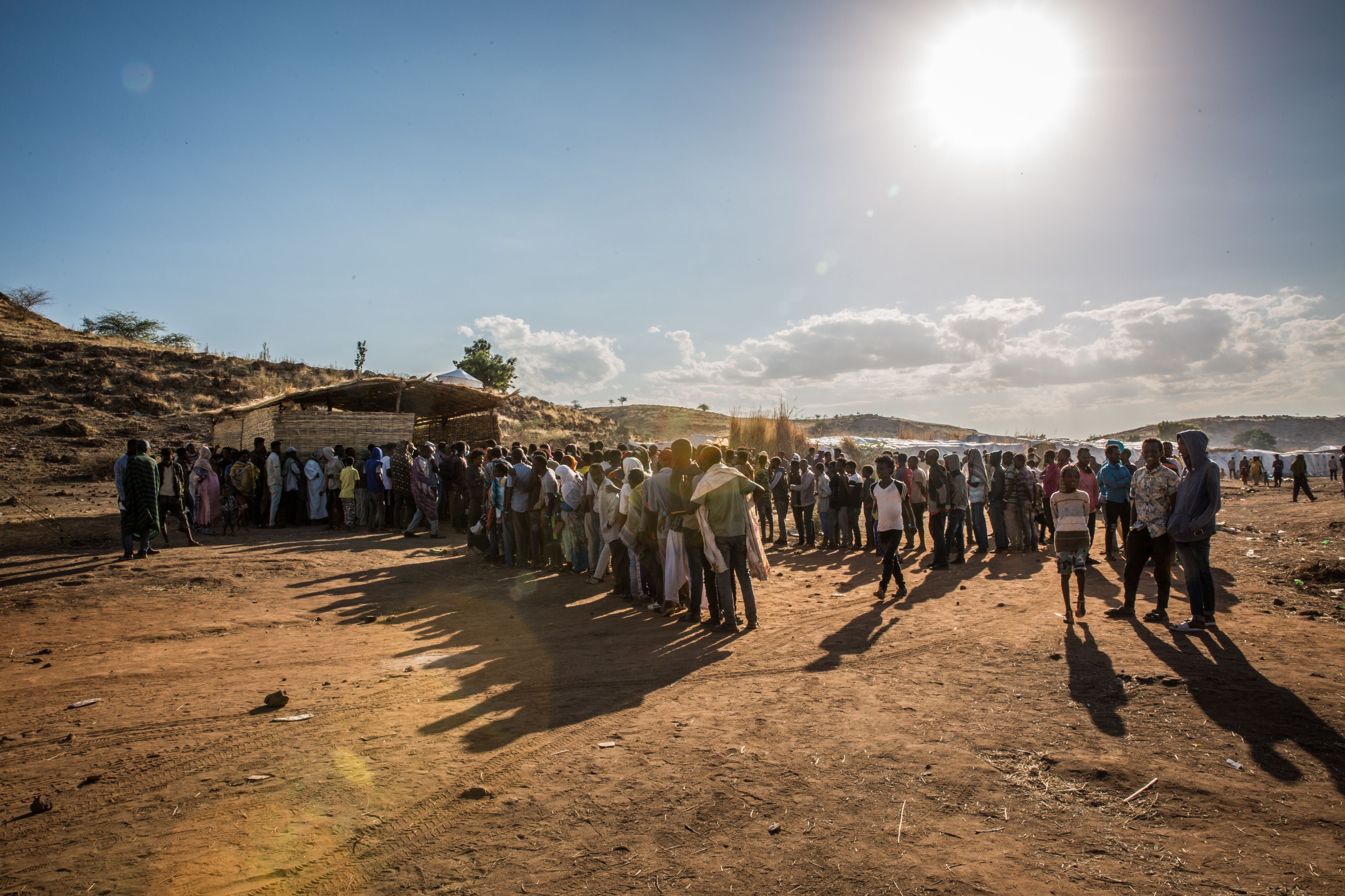 A large group of people are queuing up to register outside a refugee camp. The ground is dry and the sun is scorching.