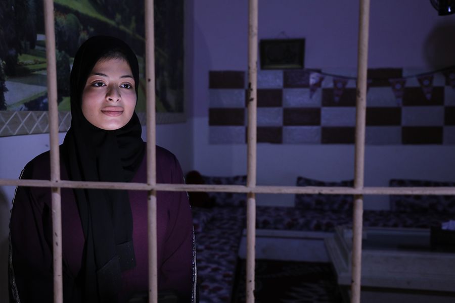 Omaia stands in a dark room. She is looking out through a barred window.