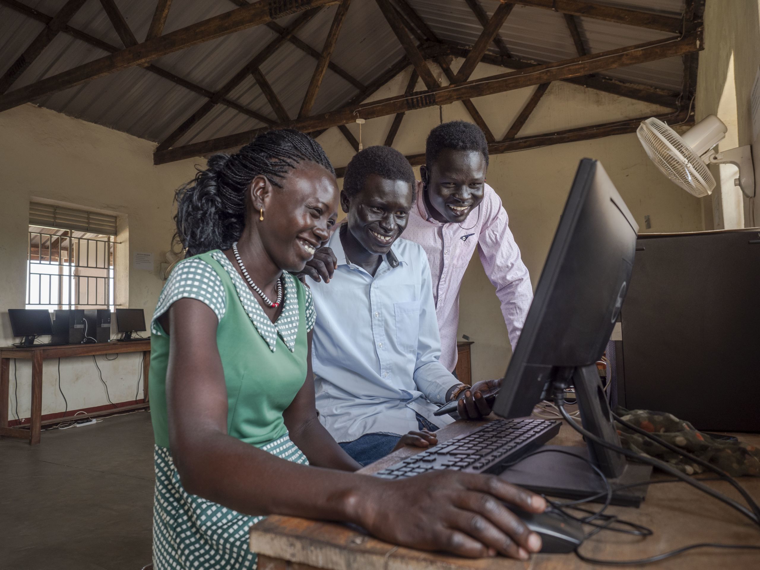Tre unge studenter, en kvinne og to menn, ler mens de leser noe på dataskjermen foran dem.